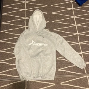 Ljusgrå hoodie från NOFS - Säljer en ljusgrå hoodie från NOFS med vit stjärnlogga och text på bröstet. Tröjan har huva med dragsko och en stor magficka. Perfekt för dig som gillar en avslappnad och stilren look.