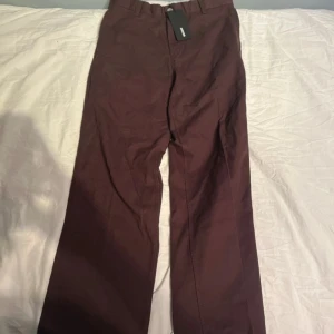 Vinröda Jeans från Weekday - Snygga vinröda chinos från Weekday med raka ben och klassisk passform. Byxorna har hällor för bälte, två bakfickor med knapp och är tillverkade i ett mjukt bomullsmaterial. Perfekta för en clean och stilren look.