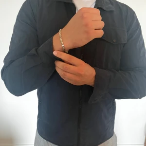 Svart overshirt från Woolrich Archive - Snygg svart overshirt från Woolrich Archive med klassisk krage, bröstficka med knapp och dragkedja framtill. Jackan är i bomull och har en clean, minimalistisk look som passar perfekt till lager-på-lager-stil. Långärmad och med diskreta fickor på sidorna.