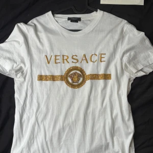 Vit Versace t-shirt Medusa - Vit t-shirt från Versace med ikoniskt guldigt Medusa-motiv och Versace-logga broderat på bröstet tiöösammans med stenar. Klassisk rund halsringning och korta ärmar. Tillverkad i mjuk bomull för en skön känsla och exklusiv look.
