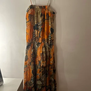 Blommig långklänning från etc. - Färgstark långklänning från etc. i 100% bomull med smala axelband. Klänningen har ett blommigt mönster i orange, beige och grönt, samt smockdetaljer upptill och i midjan. Perfekt för sommarens varma dagar och festivalvibbar.