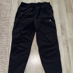 Svarta Jordan byxor med snörning - Svarta cargopants från Jordan med elastisk midja och snörning. Byxorna har dragkedjeficka på benet, justerbara benslut med snörning och en loose passform. Perfekta för en sportig och avslappnad stil.