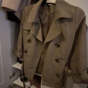 Kort beige trenchcoat från H&M - Snygg kort trenchcoat i beige från H&M med dubbelknäppning, klassisk krage och bälte i midjan. Jackan har långa ärmar och stora knappar framtill. Perfekt för dig som vill ha en stilren och tidlös look. Man kan alltid försöka pruta ner priset!