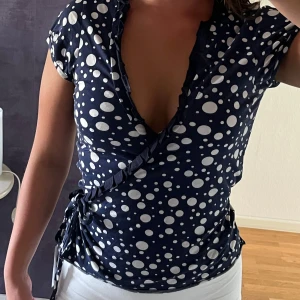 Mörkblå omlottopp med prickigt mönster - Supersnygg mörkblå omlottopp med vita prickar i olika storlekar. Djup v-ringning och knyt i sidan ger en cool och feminin look. Korta ärmar och mjukt material som känns skönt mot huden. 💞