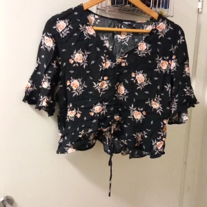 Blommig svart blus från H&M Divided - Supersöt svart blus med blommigt mönster från H&M Divided. Modellen är croppad med volangkant, korta ärmar och snörning framtill. Perfekt för dig som gillar romantiska vibes och vill ha något luftigt och snyggt till sommaren.