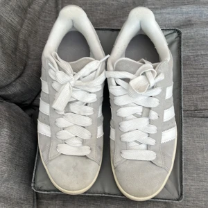 Adidas Campus grå/vita sneakers - Snygga Adidas Campus sneakers som är ljusgrå. Vita tjocka skoband. Vita detaljer på sidorna. Mycket bekväma och sköna. Perfekta skor för en avslappnad stil. 