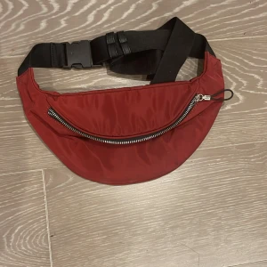 Röd magväska med dragkedja - Snygg röd magväska i syntetmaterial med justerbart svart band och silvriga dragkedjor. Perfekt för att bära mobilen, plånboken och andra smågrejer. Väskan har ett stort fack och ett mindre innerfack med dragkedja för extra säkerhet.