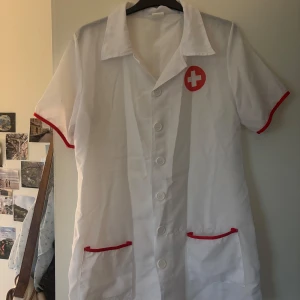Vit sjuksköterskekläning med röda detaljer - Vit sjuksköterska klänning i 100% polyester med röda detaljer. Knappar framtill och klassisk krage. Sjuksköterskeemblem på bröstet. Kommer med matchande huvudbonad med rött kors. Perfekt för halloween fest eller maskerad!!