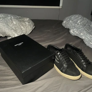 Svarta Saint Laurent sneakers - Snygga svarta sneakers från Saint Laurent med vit logga på sidan och beige platt sula. Skorna är använda i 2 veckor bara och dem har en look att de ser smutsiga ut👍