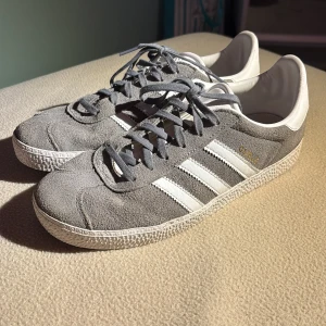 Grå Adidas Gazelle sneakers - Snygga Adidas Gazelle sneakers i grå mocka med vita detaljer och klassiska tre ränder på sidorna. Skorna har snörning, vit sula och vit häl med Adidas-logga. Perfekta för dig som gillar retrostil och vill ha något stilrent till vardags.