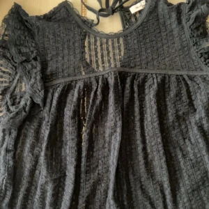 Svart blus med spets och volang - Supersöt svart blus från Nelly med transparent spets, volangdetaljer på axlarna och rund halsringning med knyt i nacken. Såå fin!