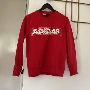 Röd Adidas sweatshirt med tryck - Röd sweatshirt från Adidas med stort vitt logotyptryck framtill och liten logga i nacken. Tröjan har rund hals, långa ärmar och ribbade muddar. Perfekt för en sportig och avslappnad stil.