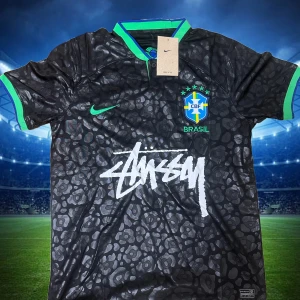 Brasil x Stüssy x Nike svart fotbollströja - Unik svart Brasilien fotbollströja i samarbete med Stüssy och Nike. Tröjan har ett subtilt leopardmönster, gröna detaljer på krage och ärmslut, samt Brasil-logga och Stüssy-tryck på bröstet. Tillverkad i lätt och ventilerande material, perfekt för match eller streetstyle.