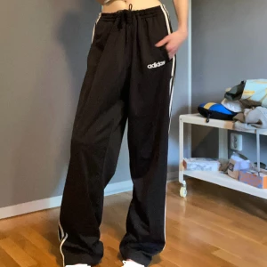 Adidas byxor - Äkta vintage från 90/00-talet! Svarta Adidas byxor med vita ränder längs sidorna och tryckt logga på låret. Vida ben & knappar längs hela sidan som kan öppnas för en cool look. Resår & snörning i midjan, passar många storlekar! Kan även ha dom på olika sätt (låg/mid/hög-waist). Perfekta för träning eller chill.😍Står strl L på lappen, midja 42cm & ner/uppåt (justerbart). Innerben 78cm. Inga defekter, bara att lilla sido märket under fickan har lossnat från sömmen lite (sista bild), går att laga!🙏🏻