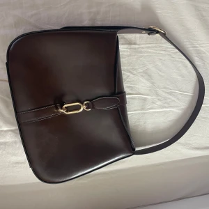 Mörkbrun axelväska i skinn med gulddetaljer - Säljer en stilren mörkbrun axelväska i slätt skinn med justerbart axelband och snygga guldfärgade metalldetaljer.  Använd få gånger. 👜👜