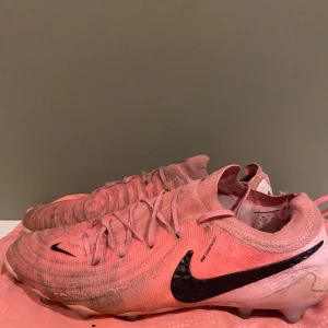Nike rosa fotbollsskor strl 45 - Säljer ett par rosa Nike phantom elit med påse  fotbollsskor med svart swoosh och snörning. Skorna har stickad ovandel och plös, samt dobbar för gräsplan. Lätta och flexibla med modern design, perfekta för dig som vill sticka ut på planen.