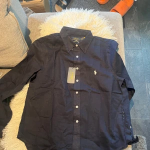 Ralph lauren linneskjorta  - Klassisk mörkblå skjorta från Polo Ralph Lauren i bomull. Skjortan har lång ärm, knappar framtill och den ikoniska broderade loggan på bröstet. Snygg krage och normal passform, perfekt för en clean och stilren look.