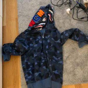 BAPE blå camo hoodie shark zip L - Säljer en ikonisk BAPE hoodie i blå camo med hajmotiv på huvan och broderade bokstäver. Tröjan har dragkedja hela vägen, känguruficka och är i mjuk bomull. Perfekt för dig som vill sticka ut med streetwear-vibe.
