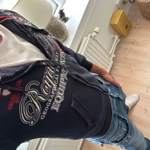  Geographical Norway Hoodie - Svart hoodie från Geographical Norway med stora broderade detaljer och tryck i vitt, beige och rött. Tröjan har huva med snörning, dragkedja framtill och känguruficka. Skriv vid funderingar 🙌