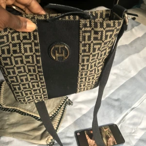 Svart och beige axelväska från Tommy Hilfiger - Snygg axelväska från Tommy Hilfiger med beige och svart mönster och stort TH-emblem framtill. Väskan har justerbar axelrem och är tillverkad i textil med detaljer i syntet. Perfekt för dig som vill ha en stilren och trendig väska med logomönster.