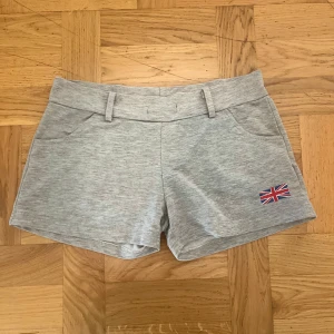 Grå shorts - Snygga ljusgrå shorts med bekväm passform och bälteshällor. Mjukt bomullsmaterial och två fickor framtill. På ena benet finns en liten Union Jack-flagga som cool detalj. Perfekta för sommardagar och chill häng.