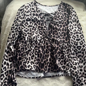 Leopardmönstrad blus med knyt - Snygg blus med leopardmönster i svart, beige och bruna toner. Modellen har långa ärmar, volang nedtill och två dekorativa knytband framtill. Materialet är glansigt och känns lätt mot huden, perfekt för dig som vill sticka ut med ett djuriskt print.