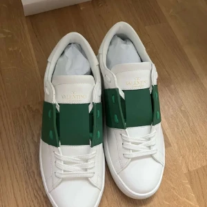 Valentino Garavani Open sneakers vit/grön - Snygga Valentino Garavani Open sneakers i vitt skinn med en bred grön läderdetalj över snörningen. Klassisk rund tå, vita snören och diskreta nitar på hälen. Perfekta för dig som vill sticka ut med stilrena och lyxiga detaljer.