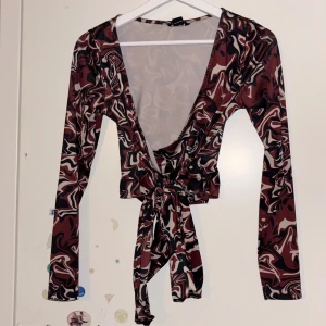 Mönstrad topp från Monki - Snygg mönstrad topp från Monki med långa ärmar och djup v-ringning. Toppen har ett abstrakt mönster i rödbruna och svarta toner och knytning framtill. Perfekt för en trendig look!