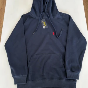 Mörkblå hoodie från Polo Ralph Lauren - Säljer en snygg mörkblå hoodie från Polo Ralph Lauren med det klassiska röda logotypen på bröstet. Tröjan har en rymlig ficka framtill och justerbar huva med dragsko. Perfekt för en avslappnad stil.