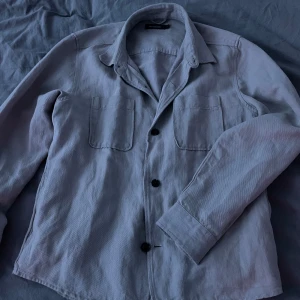 Beige overshirt från Dressmann - Säljer en stilren beige overshirt/safarijacka från Dressmann. Den har långa ärmar och knappar framtill. Perfekt för lager-på-lager-stil och passar till många olika outfits. Perfekt nu till våren🙌 modellen är 180, 60kg