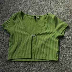Grön ribbad kortärmad topp från H&M Divided - Säljer en grön ribbad kortärmad topp från H&M Divided. Toppen har en v-ringning och en söt fjärilsdetalj framtill. Perfekt för en avslappnad stil.