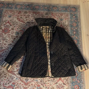 Svart quiltad jacka från Burberry Brit - Säljer en elegant svart quiltad jacka från Burberry Brit. Jackan har en klassisk krage och knappar framtill. Insidan har det ikoniska Burberry-mönstret. Perfekt för en stilren look.