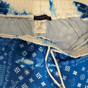 Blå mönstrade shorts från Louis Vuitton - Snygga blå shorts från Louis Vuitton med ett unikt mönster av vita moln och märkets ikoniska logotyper. Shortsen har en elastisk midja med snörning för justerbar passform. Perfekta för en avslappnad stil med en lyxig touch. (Helt oanvänd, Louis Vuitton taggen kan jag sätta tillbaka på samma plats också!)