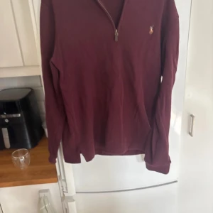 Vinröd tröja med dragkedja från Polo Ralph Lauren - Säljer en stilren vinröd tröja från Polo Ralph Lauren med dragkedja vid halsen. Tröjan har långa ärmar och är perfekt för en avslappnad stil. Den ikoniska loggan är broderad på bröstet. Perfekt för kyligare dagar!