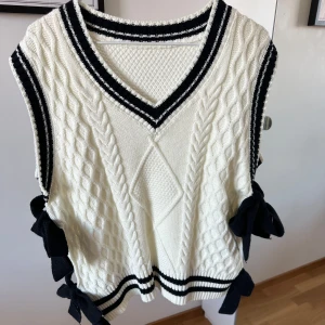 Vit stickad väst  - Snygg vit stickad väst med svarta ränder och v-ringad design. Västen har rosetter på sidorna och ett klassiskt kabelstickat mönster framtill. Den är i st M och är ifrån Cider 🫶🏼
