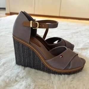 Bruna kilklackar från Tommy Hilfiger - Snygga bruna kilklackar från Tommy Hilfiger med justerbart spänne och flätad sula. Endast använda en gång.