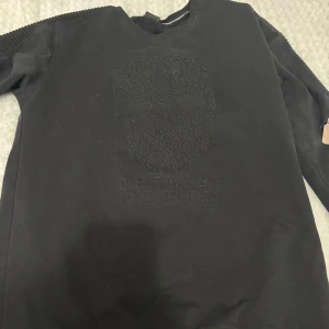 Svart tröja från Philipp Plein - Snygg svart tröja från Philipp Plein med ett coolt dödskallemönster på framsidan. Tröjan har långa ärmar och dragkedjedetalj vid ärmsluten på bara ena armen. Perfekt för en stilren look. (Pris kan diskuteras) 