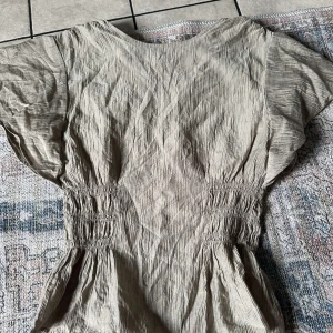 Beige tröja med korta ärmar - Säljer en stilren beige tröja med korta ärmar och smockdetaljer i midjan. Perfekt för en avslappnad look. Tröja har en luftig passform och är gjord i ett lätt material.