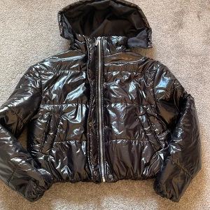 Svart glansig pufferjacka från Boohoo - Säljer en svart glansig pufferjacka från Boohoo. Jackan har en dragkedja framtill och en mysig huva. På 4 bilden ser man en liten ”rispa” på jackan dock inget man läger märket till