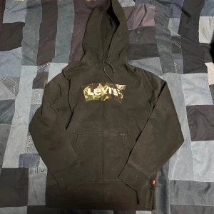 Svart hoodie från Levi's med kamouflagemönster - Säljer en svart hoodie från Levi's med en cool kamouflagemönstrad logga på bröstet. Tröjan har en klassisk känguruficka och justerbar huva. Perfekt för en avslappnad stil.