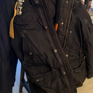 Svart parkajacka från Parajumpers - Säljer en svart parkajacka från Parajumpers med pälsdetaljer på huvan. Jackan har flera fickor och en dragkedja framtill. Den är långärmad och har en broderad logotyp på ärmen. Perfekt för kalla dagar!