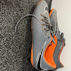 Grå och orange Nike Hypervenom fotbollsskor - Säljer ett par grå och orange Nike Hypervenom fotbollsskor med snörning. Skorna har en slimmad passform och är designade för optimal prestanda på planen. Perfekta för fotbollsspelare som söker både stil och funktion.