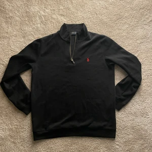 Svart tröja med dragkedja från Polo Ralph Lauren - Säljer en stilren svart tröja från Polo Ralph Lauren med dragkedja och röd logga på bröstet. Perfekt för en avslappnad men snygg look. Tröjan har långa ärmar och en hög krage.