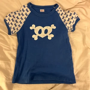 Blå baby tee med dödskallemotiv - Cool blå t-shirt från Villervalla med ett stort vitt dödskallemotiv på framsidan. Ärmarna är korta och har ett mönster av små blå dödskallar på vit bakgrund. Perfekt för en avslappnad stil!