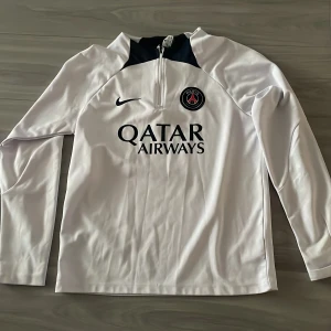 Vit långärmad fotbollströja från Nike - Säljer en vit långärmad fotbollströja från Nike med PSG-logga och Qatar Airways-tryck på framsidan. Tröjan har en dragkedja vid halsen och är tillverkad i Dri-FIT-material för optimal komfort. Perfekt för fotbollsfans! ⚽