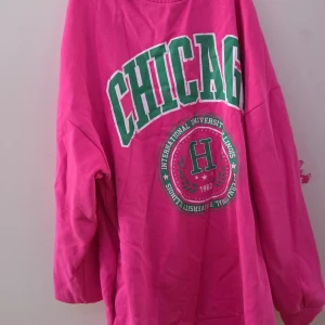 Rosa oversized tröja med tryck - Säljer en cool rosa oversized tröja med texten 'CHICAGO' i grönt och vitt. Perfekt för en avslappnad stil. Tröjan har långa ärmar och en rund halsringning. Storleken är xxs men skulle säga att den passar xxs-s