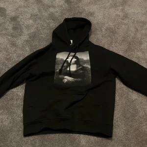 Svart hoodie med Mona Lisa-tryck - Säljer en svart hoodie med ett unikt Mona Lisa-tryck där ögonen är täckta. Texten 'Florence, 1503' syns ovanför motivet. Perfekt för dig som gillar konst och streetwear. Hoodien har en klassisk passform med långa ärmar och en bekväm huva.