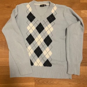 Ljusblå stickad tröja med argylemönster - Snygg ljusblå stickad tröja från Noble med klassiskt argylemönster i svart och vitt. Tröjan har en v-ringad hals och långa ärmar med ribbade muddar. Perfekt för en stilren look!