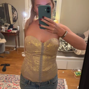 Guldig korsettopp - Säljer en elegant guldig korsettopp med blommigt mönster. Toppen är axelbandslös och har en smickrande passform med spännen framtill. Storleken är 42 men passar mig som har 38!! Perfekt för en festlig kväll!