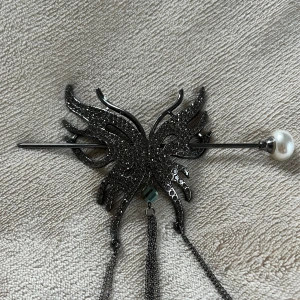 Hårnål med fjärilsdesign och pärldetalj - Elegant hårnål i metall med en fjärilsformad design. Den har en glittrig yta och en pärla i ena änden. Från fjärilen hänger flera kedjor som ger en extra touch av stil.